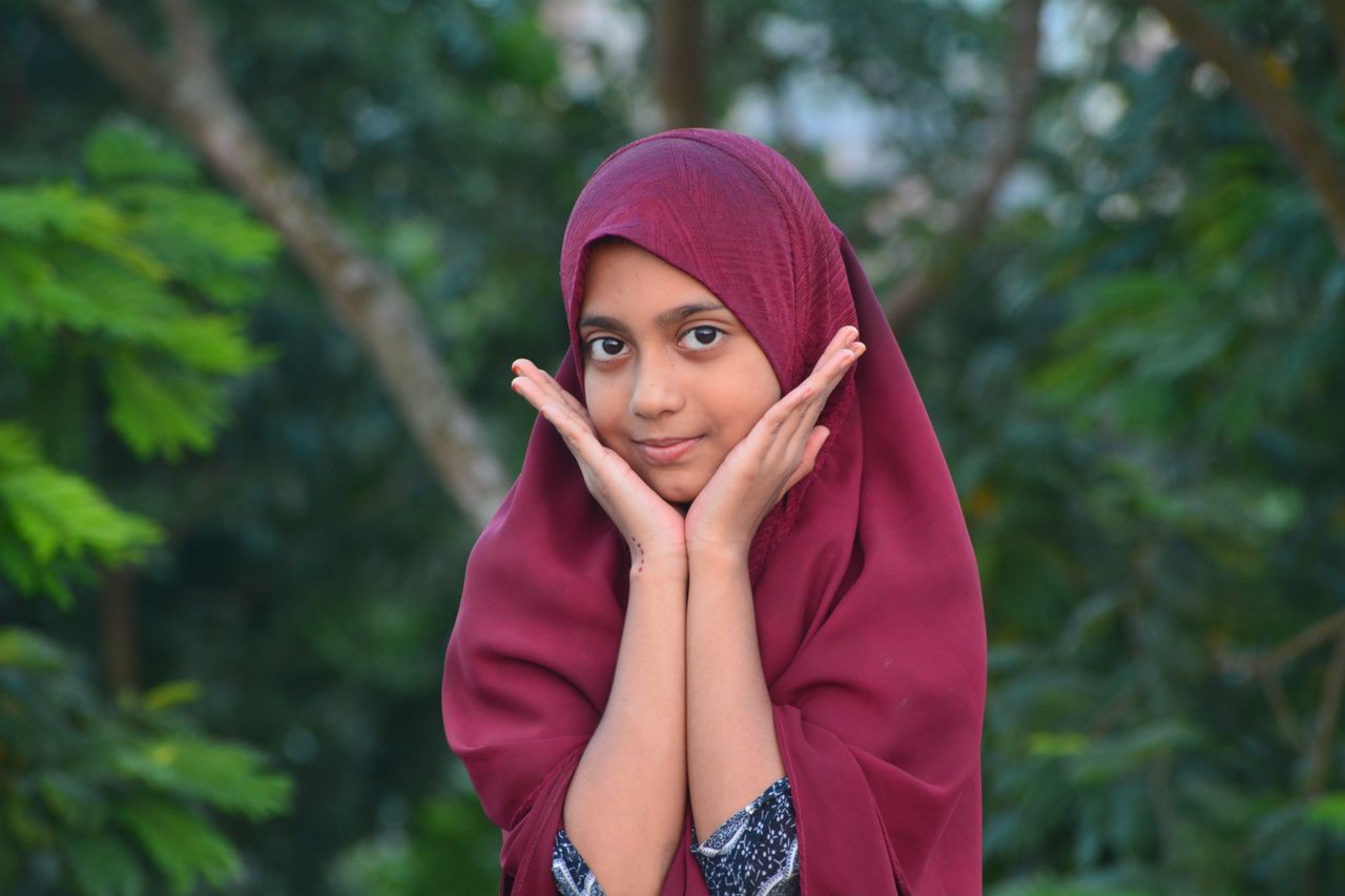 Portrait of a young girl wearing a hijab, hands on cheeks, outdoors.